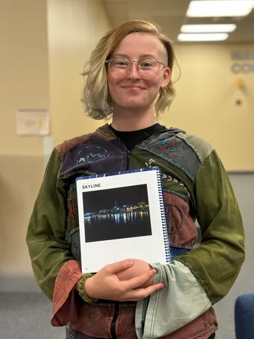 Bella stands, smiling at the camera and holding her submission to the 2024 Totem Literary Magazine