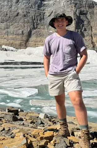 Cooper Cernuska stands in front of a river and wearing a hat and smiling