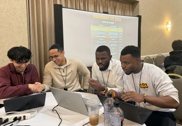 a group of four students working on laptops, two are shaking hands and smiling. In the background the points standing shows the ���Ǻ���hacks.Club team is currently in first place.