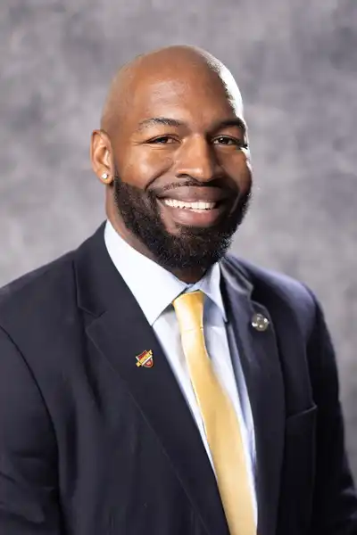 A'Darius smiles, head tilted to the left in a black suit for a headshot on a gray background