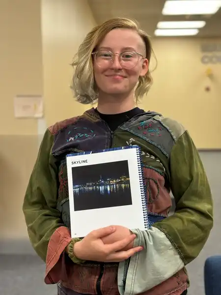Bella stands, smiling at the camera and holding her submission to the 2024 Totem Literary Magazine