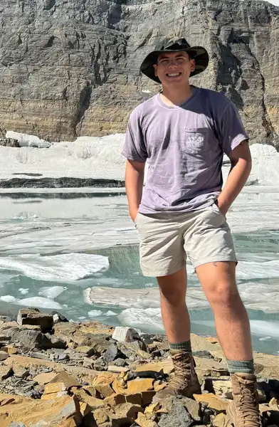 Cooper Cernuska stands in front of a river and wearing a hat and smiling