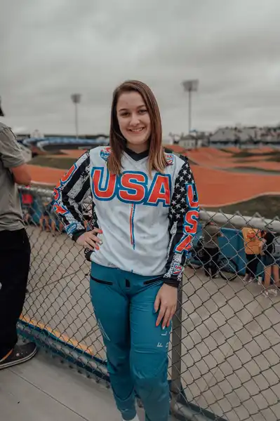 Kady Brink stands with one hand on her hip in front of the BMX track in Rockhill, South Carolina. She is wearing her USA jersey and smiling.