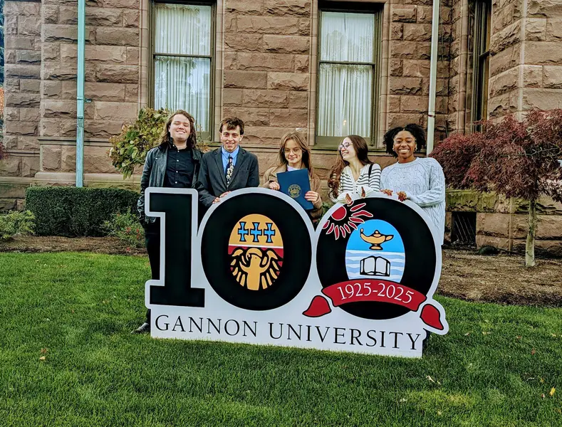 Five students stand behind the ������Ƶ centennial sign in front of Old Main. In the center, Cadence Hoover holds a proclamation from the Mayor of the City of Erie.